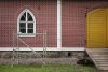 Punatiilen näköiseksi maalattua seinää, jossa ikkuna ja keltainen kaksoisovi. Ikkunan edustalla rakennusteline.